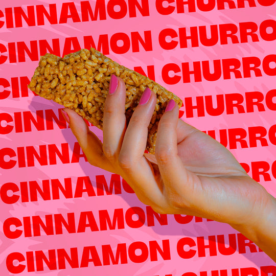 Hand holding a Wild Fox Cinnamon Churro Protein Bar with red 'Cinnamon Churro' text on a pink background