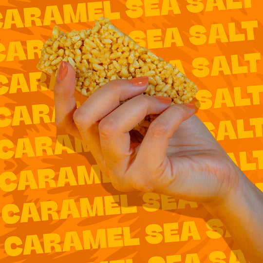 Hand holding a Wild Fox Caramel Sea Salt Protein Bar on an orange background with 'Caramel Sea Salt' text.