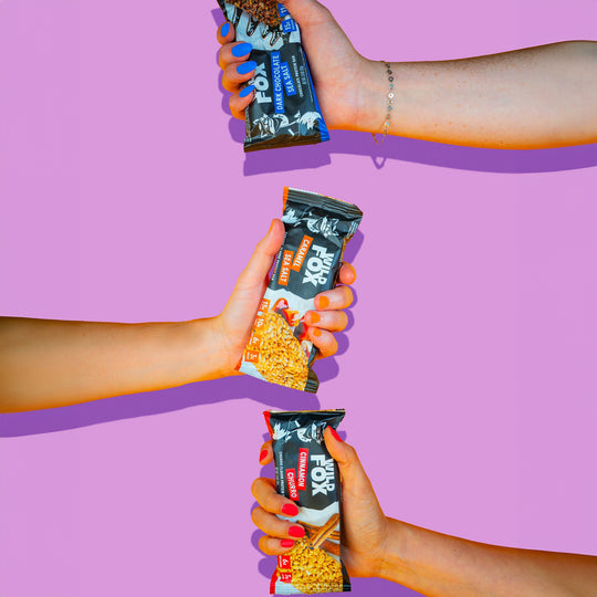 Three hands holding different flavors of a snack bar against a purple background